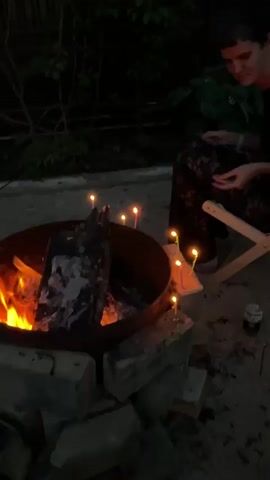 A video shows a person sitting in a chair next to a fire pit at night. The fire pit contains burning logs and several lit candles. The person is smiling and holding something in their hands. The background is dark with some foliage visible.