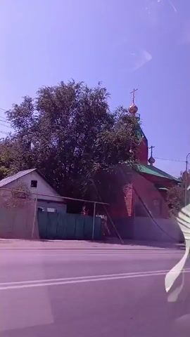 The video shows an exterior view of a church. The church is red with a green roof and a golden dome topped with a cross. Trees and other buildings are visible around the church. Cars are driving past on a road in front of the church. The sky is clear and blue.