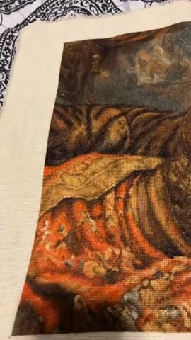 This is a close-up video of a nearly finished cross-stitch tapestry. The tapestry depicts a woman with dark hair, wearing elaborate gold jewelry and a red and orange patterned headscarf. She is looking towards the viewer with blue eyes and a slight smile. Her right hand is resting on her chin, and her left arm is bent. Next to her is a tiger, its head and part of its body visible. The background appears to be a rocky or desert-like landscape. The tapestry is displayed on a light-colored surface, possibly a table or easel, and a small portion of a black and white patterned fabric is visible at the top of the frame. The stitches are clearly visible, creating a detailed and textured image.
