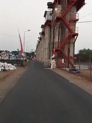 The video shows a view from a vehicle driving on a paved road alongside a large concrete structure with red metal stairs. The concrete structure appears to be a dam or a similar industrial facility, with a series of large vertical pillars. Red flags on poles and white sacks are placed near the base of the structure. In the distance, several motorcycles are visible, suggesting other vehicles are also present. The sky is overcast.