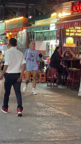 The scene unfolds on a bustling street, likely during the evening or at night, evidenced by the presence of neon signs and the general lighting conditions. A woman in a light blue t-shirt featuring a cartoon bear, denim shorts, and sneakers walks directly towards the camera. She carries a white shoulder bag. To her right, a couple walks with their arms around each other. The man wears a white patterned shirt and blue shorts, while the woman is in a green shirt and white shorts. Behind them, people are seated at the Lucky Bar, which displays a neon sign with the text "we only want to get drunk." Additional people can be seen seated at tables along the street. Overall, the setting appears to be a lively, tourist-oriented area.