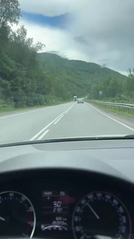 The video is filmed from the perspective of a driver inside a car, showing the dashboard and the road ahead. The dashboard displays a speedometer with the needle indicating approximately 90, and an odometer showing 6222 km. The road is a two-lane highway that curves gently to the right, with mountains covered in lush green trees on either side. The sky is overcast with some clouds. Several vehicles are visible on the road, including a white truck, a few cars, and a large semi-trailer truck. One car in the foreground on the left appears to have damage to its front bumper. The video captures the car moving along the highway at a steady pace.