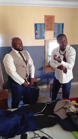 The video shows two men in a room. The man on the left is bald, wearing a white shirt, brown vest, dark slacks, and a tie. He’s smiling. The man on the right is wearing a white shirt, dark slacks, and a tie. He’s fastening his cuff. Between them is a table with various items, including what looks like clothing and other objects. Behind the men is a wall with light yellow paint on the top and grey paint on the bottom. There are two framed pictures hanging on the wall.