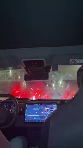 The video shows the interior of a car from the perspective of the passenger seat. In the foreground, a large touchscreen display is visible, showing what appears to be a music player with album art. The driver's side of the car is partially visible on the left, including the steering wheel. Through the windshield, a line of cars is stopped, indicated by multiple red taillights. The environment appears to be a tunnel or an enclosed roadway, with overhead lighting and barriers. There is some condensation or mist on the windshield. The overall scene suggests the car is stuck in traffic.