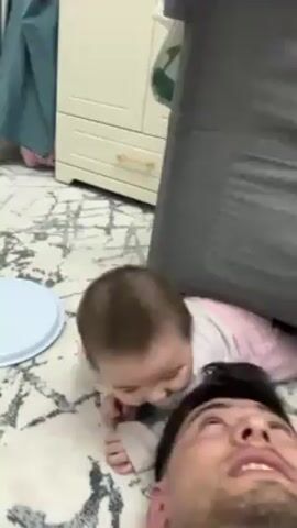 A father is lying on his back on a rug with a baby girl crawling on top of him, bumping her forehead on his forehead. The baby has light brown hair and is wearing a light beige shirt and pink pants. The father has dark hair and a beard, and he is smiling. Next to the father is a gray laundry hamper. The rug has a gray and white pattern. Behind them, there is a white dresser with gold handles. The video is shot from a low angle, looking up at the father and baby.