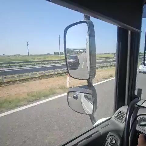 The video shows the view from the driver's seat of a large truck driving on a highway. The driver's side mirror is visible in the foreground, reflecting the road behind. The truck is in the right lane of a multi-lane highway. In the distance, other vehicles can be seen, including cars and trucks. The landscape outside the highway is flat and rural, with fields and some trees. The sky is clear and blue. The video appears to be a screen recording of a dashcam feed.