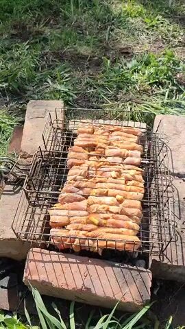 The video shows raw sausages grilling on a square metal grate. The grate is supported by bricks, forming a makeshift barbecue. The ground around the barbecue is grassy. Smoke rises from the sausages as they cook.