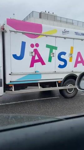 The video captures an ice cream truck driving down a wet street. The truck is white with colorful lettering that appears to say "JÄTSABI". It also has images of ice cream and the phone number "TELLIMINE: 5331 7001". The truck is passing a modern-looking apartment building with balconies and a dark-colored car parked on the side of the road. The street is asphalt and appears to be a residential area. The weather is overcast.