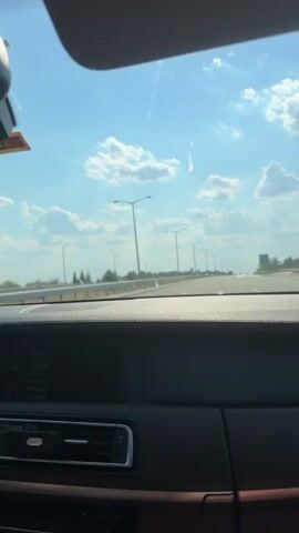 This media file shows a view from inside a car, looking out the windshield. The **scene** is a highway with a clear blue sky dotted with white, puffy clouds. The **setting** appears to be a sunny day, possibly late morning or early afternoon, given the bright sunlight.

The **subjects** are primarily the highway and its surroundings. There are rows of streetlights lining the road, suggesting a well-developed infrastructure. Beyond the highway, there are trees and some structures, including what appears to be a green road sign in the distance on the right. The car's dashboard is visible in the foreground, with a dark, glossy finish. A digital display screen on the dashboard shows some text, which appears to be a playlist or song list. The visible text on the screen includes words like "BROTHERS," "REAKTION," "RO AD," "NO FEAR," "REALITY," "RYTHMOS," and "ZADORA."

The **activity** taking place is travel by car. The perspective suggests the car is in motion, moving along the highway. There are no people clearly visible, but the viewpoint implies someone is driving or a passenger. The **weather** is fair and sunny, contributing to a pleasant driving experience. The **location cues**, such as the streetlights and the type of highway, are consistent with a modern road system. There is no explicit indication of emotion, but the bright weather and open road could evoke a sense of freedom or journey. The location context of Havsa, Turkey, places this scene in a specific geographical area.