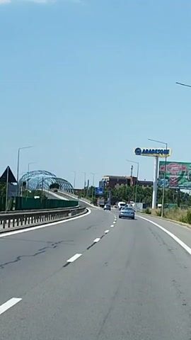 The video shows a view from a vehicle traveling on a multi-lane highway. The road has white lane markings. On the left side of the road, there is a green sound barrier and a guardrail. Ahead, a large, futuristic-looking pedestrian bridge with a curved, transparent roof spans over the highway. Cars are visible on the highway, some in front of and some in the opposite direction. On the right side of the highway, there are streetlights, a billboard for "ARADEION", and some buildings. The sky is clear and blue.