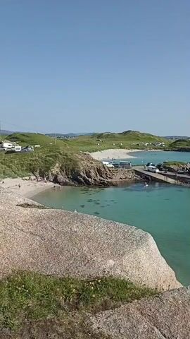 The video shows a panoramic view of a coastal area on a clear, sunny day. The foreground is dominated by a large, weathered granite rock formation. Beyond this, the turquoise water of the sea is visible, with several small boats moored and others sailing or being rowed. A stone pier extends into the water, with vehicles, including white vans and what appear to be campers or caravans, parked on it. People are seen on the pier and on a sandy beach to the left. The background features rolling green hills dotted with more buildings and what appear to be more campers or tents. The horizon is a clear blue sky. The overall impression is of a peaceful, scenic seaside location that is actively being used for recreation and possibly as a small ferry or transport hub.