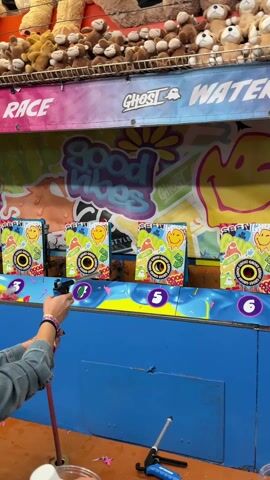 The video shows a water race game being played at a carnival. A person is standing at a water gun station, aiming at a target. There are four stations in total, with numbers 3 through 6 indicating the spots. Above the target are various stuffed animals arranged in a row. The game is called "Ghost Water Race," and it has a colorful background with words like "Good Vibes" and smiley faces. In the background, there are other players and carnival-goers. At the beginning of the clip, the first player shoots at the target, popping the balloon over the target. The second player then shoots at another target, making the balloon pop over the corresponding target.