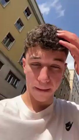 A fair-skinned young man with curly dark hair is filming himself with a camera, probably a phone, on a bright sunny day. He is wearing a white Nike t-shirt. He is outside on a city street with light colored buildings in the background, some with awnings over outdoor tables. He is looking at the camera and moving it to show the scenery behind him. The sky is blue and sunny with a few white clouds.