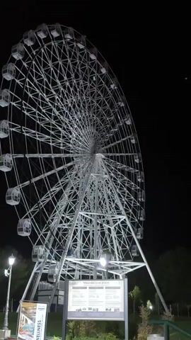 A static shot depicts a large, white Ferris wheel at night. The wheel is brightly lit and contrasts sharply against the dark sky. The wheel's structure is visible, consisting of a network of metal beams and enclosed gondolas. Below the wheel, there is a sign with the text "Mineralno jezero Jezero" visible, and some greenery. A lamp post illuminates the foreground on the left. The environment appears to be a park or public space.