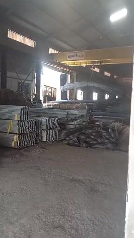 The video shows the interior of a warehouse filled with various metal materials. In the foreground, neatly stacked bundles of metal rods or pipes are visible, secured with yellow straps. To the right, piles of coiled dark metal wire are present. In the background, a yellow overhead crane is actively lifting a bundle of long metal beams. Sunlight streams in from windows in the upper part of the warehouse, illuminating the space. A person can be seen near the stacked metal rods, appearing to be working. The floor is a rough, gravel-like surface. The overall impression is of a storage or processing facility for metal goods.