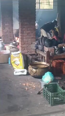 The video shows a busy kitchen scene, likely a communal cooking area or an outdoor eatery. Several people are present and engaged in food preparation. In the foreground, a large brass pot sits on the ground, suggesting it's used for cooking. To the left, a large bag of what appears to be rice or flour stands upright. Smoke billows from cooking fires in the background, indicating active cooking is taking place. People are seen tending to the fires and pots. The environment is rustic, with brick pillars, wooden structures, and various cooking utensils and containers scattered around. The lighting appears natural, with some overhead illumination. The overall impression is one of a functional, albeit somewhat cluttered, cooking operation.