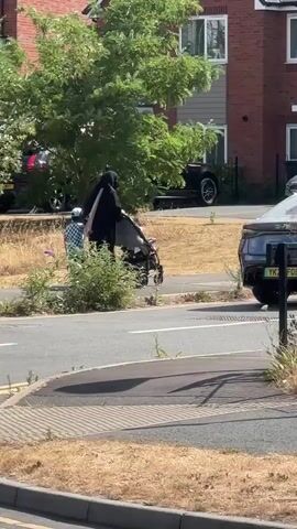 A person wearing a black niqab and a child wearing a checkered shirt are walking along a sidewalk. The person is pushing a stroller. The child is walking beside the person. The scene takes place on a sunny day. There are houses and cars in the background.