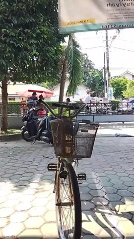 The video pans across a street scene. In the foreground, a bicycle with a basket is parked on a cobblestone path. To the right, a motorcycle is parked. Behind the motorcycle, more parked motorcycles are visible. In the background, there are buildings, trees, and street signs. A few people are walking in the background, and a car is parked on the street. The overall atmosphere appears to be a busy outdoor area, possibly a marketplace or street vendors.