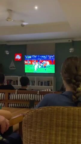 A group of people are sitting in a room and watching a soccer game on a large television screen. The room is dimly lit, with a projector mounted on the ceiling. The television is displaying a game with players in red and white uniforms. There are several rows of seating, with people sitting on couches and chairs. The walls of the room are painted green, and there are shelves filled with books behind the seating. There is also a small whiteboard with a picture of a sailboat on it. The people in the video are facing the television and appear to be engaged in watching the game.
