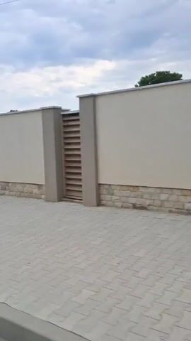 The video pans from right to left, showing a street view. In the foreground, there is a gray paved sidewalk with white lines marking the edge. A lamppost with election posters featuring a man's face is visible on the right side of the sidewalk. The lamppost is wrapped with posters from top to bottom. Behind the sidewalk, there is a wall made of light-colored stone and some horizontal wooden slats. Further back, there are some green bushes and trees, as well as the tops of buildings. The sky is overcast. The video captures a residential or commercial area.
