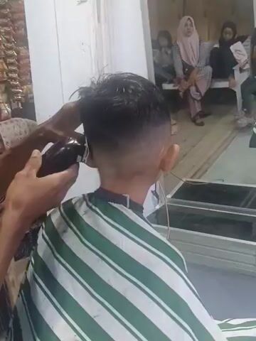 Here's a description of the video:

The video shows a boy getting a haircut in what appears to be a barbershop. The boy is seated and wearing a striped barber's cape. A barber is using electric clippers to cut the hair at the back of his head, creating a fade effect. A mirror in front of the boy reflects the barber working and shows a group of women and children sitting and watching in the background. One woman is wearing a hijab. The environment seems to be a simple, modestly decorated barbershop.