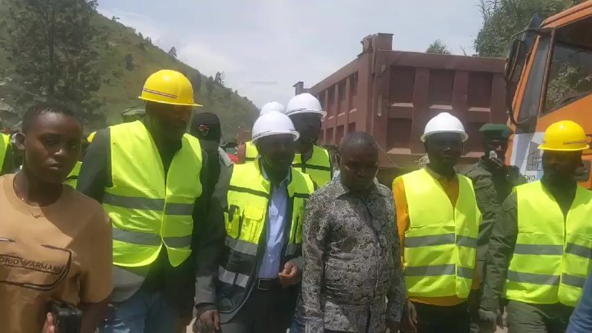 The video shows a group of approximately 10 people standing outdoors during daylight hours. Several of them are wearing hard hats, either white or yellow, and bright yellow reflective vests, suggesting a construction or engineering environment.  A large orange dump truck is parked in the background. Some of the people appear to be in conversation. The location appears to be on or near a road in a hilly or mountainous area. One individual is wearing a military-style uniform in the background, suggesting a security or official presence.