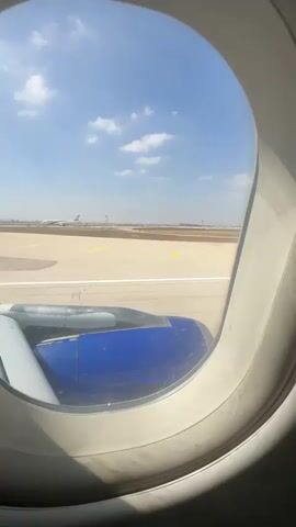 The video shows a view from an airplane window as the plane taxis on the runway. The passenger is seated on the left side of the plane, as evidenced by the engine cowling being on the left. The engine is blue. The runway is visible, with yellow markings on it. The sky is blue with scattered white clouds. In the distance, there are airport buildings and possibly other aircraft. The plane appears to be moving slowly. The sun is casting a shadow on the window frame.
