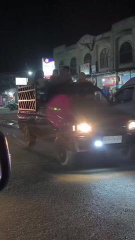 The video shows a black pickup truck driving at night in Tingkeummanyang, Indonesia. Several people are riding in the open bed of the truck. The truck has a metal cage-like structure at the back, and some of the passengers are sitting on top of it. The scene is dimly lit, suggesting it is nighttime. The truck's headlights and taillights are visible, and there are streetlights in the background. The buildings in the background appear to be illuminated, and some have arched windows. The road surface is visible, and it appears to be paved. The overall atmosphere is casual, with people enjoying a ride in the back of the truck. The license plate of the truck is visible and reads "B 9232 ZV". The weather appears to be clear.