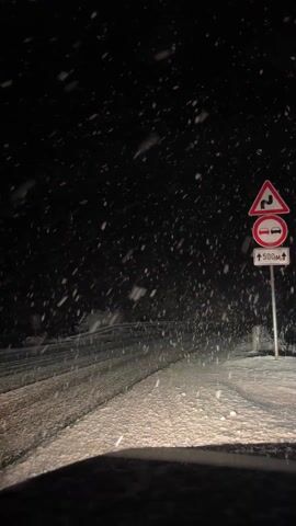 The video shows a nighttime view of a snowy road from inside a vehicle. Snow is falling heavily. A road sign on the right side of the road indicates winding roads and a no-passing zone, with the distance indicated as 500 meters. The road is covered in snow and the headlights of the vehicle illuminate the immediate area. The background is dark, with minimal visible features other than the falling snow.
