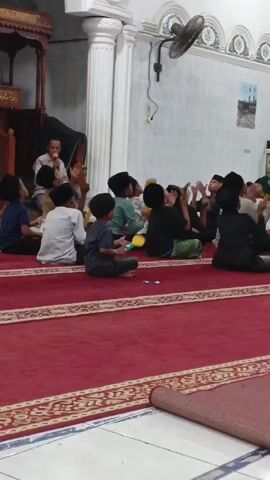 In a dimly lit room, possibly a mosque, a group of young boys are sitting on a red carpeted floor, facing a man who appears to be a teacher. The teacher is standing and speaking into a microphone, likely giving a lesson or leading a song. The boys are dressed in traditional Islamic attire, including skullcaps. Some of the boys are holding yellow toys, possibly maracas or small instruments, and seem to be engaged in a musical activity. The room has white walls with decorative patterns, a ceiling fan, and a wooden podium. The overall atmosphere suggests a religious or educational gathering for children.