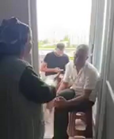 The video captures a candid moment with at least three individuals visible. In the foreground, a woman wearing a dark headscarf and a dark green vest is facing away from the camera, engaged in conversation with a man seated on a stool. The man, wearing a light-colored shirt, is looking towards the woman and appears to be speaking. Behind them, a younger person in a black t-shirt and light-colored shorts is seated, looking down at a mobile phone. The scene appears to be an outdoor or semi-outdoor setting, possibly a balcony or porch, with a bright, overcast sky. Trees and buildings are faintly visible in the background, suggesting a residential area. The overall mood seems relaxed and informal, indicative of a casual gathering or a visit. The lighting suggests daytime, but the overcast sky tones down any harsh sunlight. No text is visible in the video. The setting, with its open doorway leading to an outdoor space and the casual attire of the individuals, hints at a warm climate. The interaction between the woman and the man suggests a personal conversation, while the younger person's focus on their phone indicates a common modern social dynamic.