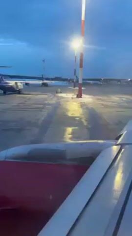 The view from inside a plane shows the airport tarmac and the wing of the plane in the foreground. In the distance, a smaller plane with a purple tail is parked on the tarmac, with orange cones nearby. The sky is a cloudy blue, and the tarmac is wet and reflecting the lights of the tall red and white striped poles along the edges of the runway. There is some snow visible on the ground on the edges of the tarmac.  The view is static.