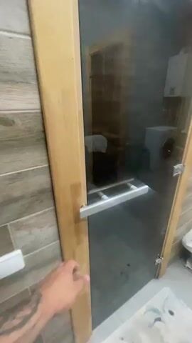 The video begins with a close-up of a person's tattooed arm reaching for a silver handle on a wooden and glass door. The camera then pans to reveal a modern bathroom with grey tiled walls and a light-colored floor. A glass-doored wooden sauna is visible in the background. In the foreground, a toddler, naked and with wet curly hair, is sitting on the floor, playing with various bath toys, including plastic vegetables and a small green truck. A showerhead and a yellow bath mat are also on the floor. A radiator with white fins is mounted on the wall beneath a tall window. The overall atmosphere suggests a warm, indoor setting, likely a bathroom or a spa area, during daytime. The toddler appears engaged and content with their playtime. There is no visible text in the video.