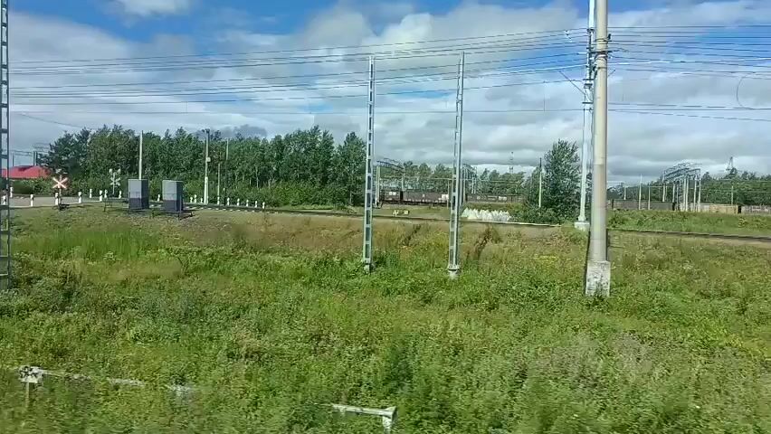 The video shows a view from a moving train on a sunny day with a blue sky and scattered clouds. The train is passing through a rural or semi-rural area with green vegetation, trees, and power lines. There are several railway signals and poles visible along the tracks. In the distance, a few buildings, including a red-roofed house, can be seen. The train tracks themselves are also visible, running alongside the train. There are no people or animals clearly visible in the frame. The video captures the passing scenery as the train travels.