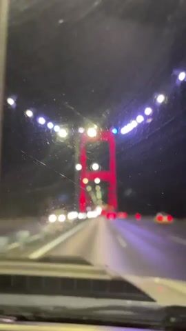 The video shows a view from the inside of a vehicle looking forward. The vehicle is on a bridge at night. The bridge is lit with string lights along the top. A large red structure, possibly a bridge tower, is visible in the center of the frame. There are headlights and taillights of other vehicles on the bridge. The road surface is visible, with lane markings. The windshield of the vehicle appears to have some smudges or debris on it.