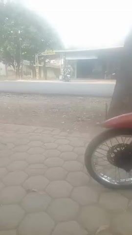 The video shows a dusty road in what appears to be a rural or semi-rural area. The footage is blurry and hazy, suggesting either poor camera quality, atmospheric conditions like dust or fog, or a combination of both.

At the beginning of the video, the camera is focused on the ground, which is a mixture of dirt, gravel, and some paving stones in the foreground.

As the video progresses, a person on a motorcycle comes into view. The rider appears to be wearing a checkered shirt. They are moving along the road.

Further into the video, a red and white vehicle, possibly a small bus or van, passes by. The specific type of vehicle is difficult to discern due to the blurriness.

The background shows glimpses of buildings, trees, and vegetation, typical of a roadside scene. There are also some utility poles visible. The overall impression is that of a roadside environment with some passing traffic. The dominant characteristic of the scene is the hazy and dusty quality of the image.
