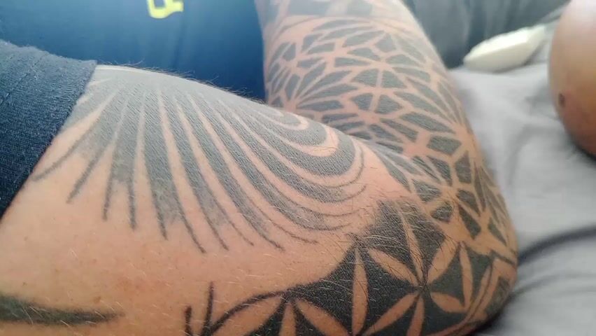 The video shows a person's tattooed arm resting on a gray surface, likely a bed. The tattoo consists of geometric patterns, including a flower-like design and lines resembling sun rays. The person is wearing a dark blue shirt with a yellow logo. In the background, there is a white object and a round, blurred object, possibly a person's head. The lighting is soft and natural, suggesting the video was taken indoors.