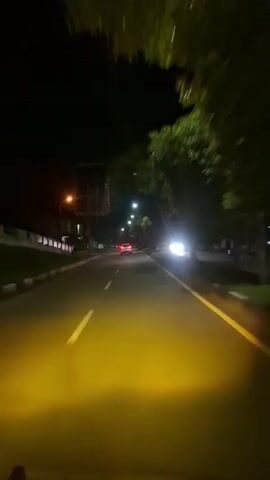 The video shows a street scene at night, filmed from the perspective of a moving vehicle. The road has yellow lines painted on it, and streetlights illuminate the scene. There are trees lining the side of the road, and some buildings and a billboard are visible in the background. A few other vehicles are on the road, including a red car ahead and a car with its headlights on approaching from the right. The scene is dark and somewhat blurry due to the movement of the camera.
