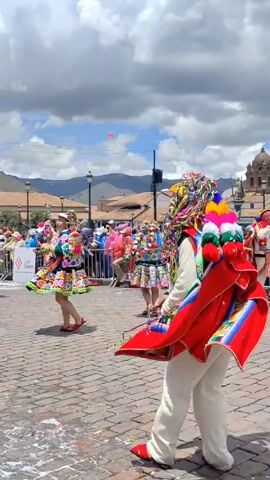 A video shows a group of people dancing in a cobblestone square, with a large church in the background. The dancers are wearing colorful traditional clothing, including hats, skirts, shawls, and vests. The video pans to follow the dancers as they move. There is a crowd of people watching the performance. The sky is cloudy with patches of blue. The square is littered with confetti.