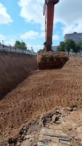 The video shows an excavator at a construction site. The excavator is digging a hole in the ground. The sky is blue with some clouds. There are buildings and trees in the background.