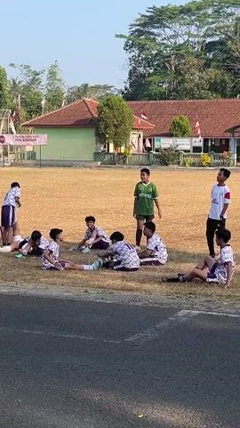 A group of young boys in soccer uniforms are gathered on a dry, grassy field in front of a school building. Some of the boys are sitting on the ground, stretching, while others are standing. A man on a motorcycle, pulling a cooler cart, drives past the field. The scene appears to be outdoors during the daytime.