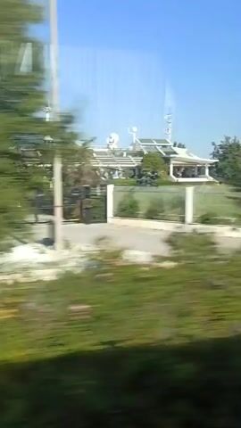 The video shows a blurry outdoor scene, likely from a moving vehicle. In the foreground, there is green grass and foliage. In the background, there is a large structure with cranes and what appears to be a ship or industrial equipment. The sky is blue and clear. The footage is shaky and out of focus, suggesting it was recorded while the camera was in motion.