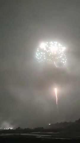 A fireworks display is happening at night. The sky is dark and cloudy. Several fireworks have exploded, creating bright bursts of light. One firework is ascending, leaving a trail of smoke. In the distance, there are lights from a town or city. The ground is dark, possibly with trees or a hill visible.