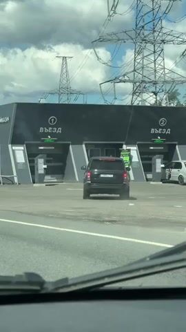 A black SUV is driving towards a car wash with two bays labeled "ВЪЕЗД" meaning "entrance." The car wash is a modern building with a black facade. Large electrical towers and power lines are visible in the background against a cloudy sky. A white SUV is parked to the right of the car wash entrance, and another white car is visible in the distance further to the right. The camera appears to be inside another vehicle, looking out through the windshield.