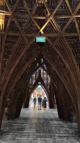 The video shows an interior view of a building with a complex bamboo structure. The structure forms a series of arches leading towards a brightly lit opening at the end. Three people are walking away from the camera, further into the bamboo tunnel. There's an exit sign visible in the center of the bamboo structure. As the camera pans upwards, the intricate geometric pattern of the bamboo construction is revealed, culminating in a dome-like ceiling with light shining through some of the openings. The overall impression is one of architectural ingenuity and complex construction.