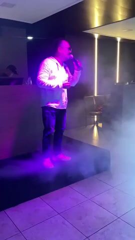 A man stands on a raised platform, holding a microphone and singing. He is wearing a white hoodie and jeans, with sunglasses on. Blue and white smoke effects swirl around him, illuminated by stage lighting. In the background, a DJ booth is visible, and other people are present in the dimly lit room, with some dancing under colored lights. The overall atmosphere suggests a musical performance or party setting.