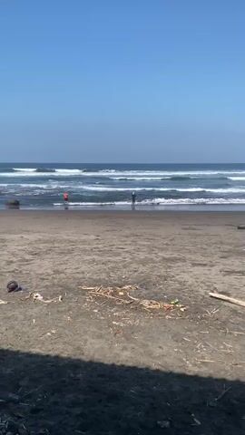 The video shows a beach scene. In the foreground, the sandy beach is littered with debris including wood, plastic, and trash. In the middle ground, two people are standing in the shallow water at the edge of the ocean. One person is wearing an orange shirt and the other is wearing dark clothing and a hat. In the background, the ocean has several waves breaking and moving towards the shore. The sky is clear and blue. The camera is static.