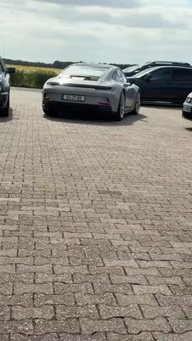 A grey Porsche 911 GT3 turns left in a paved lot, driving past other parked cars. The lot is next to a field under a partly cloudy sky.
