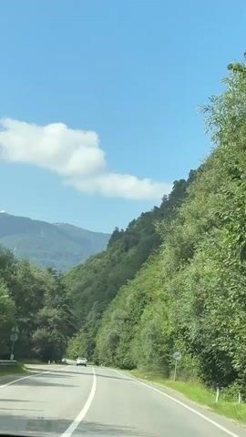 The video shows a view from a car driving on a paved road through a mountainous, forested area. The sky is blue with some white clouds. The road has white lane markings. A white car is visible driving ahead in the same direction. The car passes a sign on the right side of the road. The driver comments on the car's maneuver, saying it "changed lanes on the go."