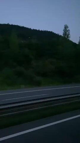 The video captures a view from a moving vehicle on a highway at dusk or night. The sky is a deep blue-grey. To the left, a densely forested hillside rises steeply. A guardrail separates the highway lanes from the embankment. The highway itself has white lane markings. In the second frame, a white car with its headlights on is visible in the distance, blurred due to motion. The rest of the video shows the same scene without the car. The overall impression is one of a journey through a dark, wooded landscape.